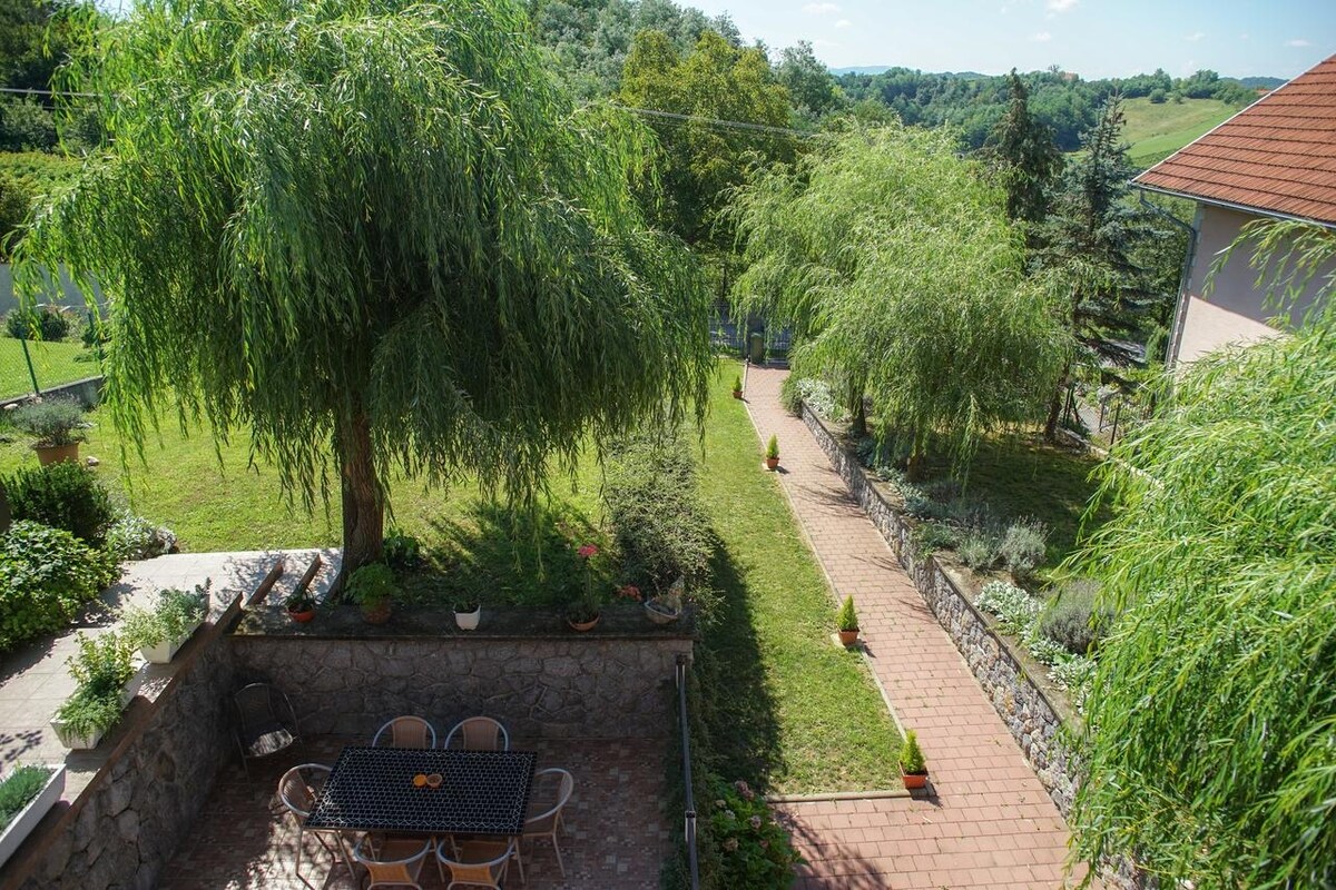 A well-maintained garden area features a stone patio with a large table and chairs, surrounded by greenery. A pathway leads through the vibrant landscape, bordered by trees and flower pots, promoting a serene outdoor space ideal for relaxation.