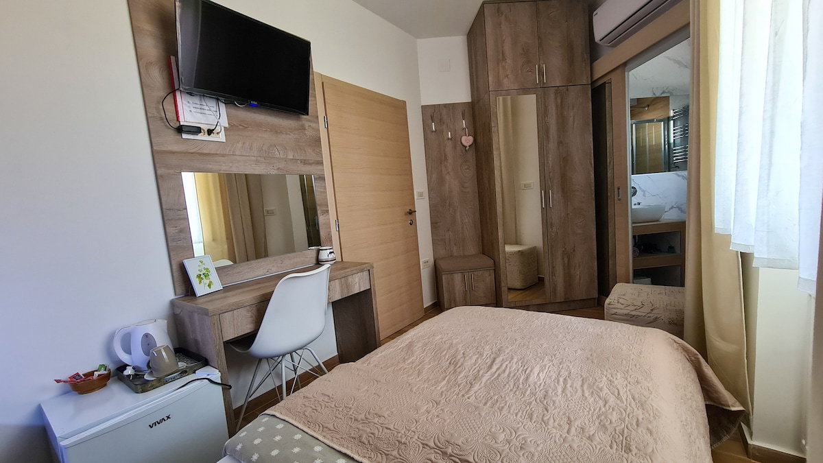 A modest bedroom is equipped with a spacious bed covered by a beige blanket. A desk with a white chair and a mounted TV is visible. Natural light filters through sheer curtains, illuminating the room. A small refrigerator and mirror are placed within the scene.