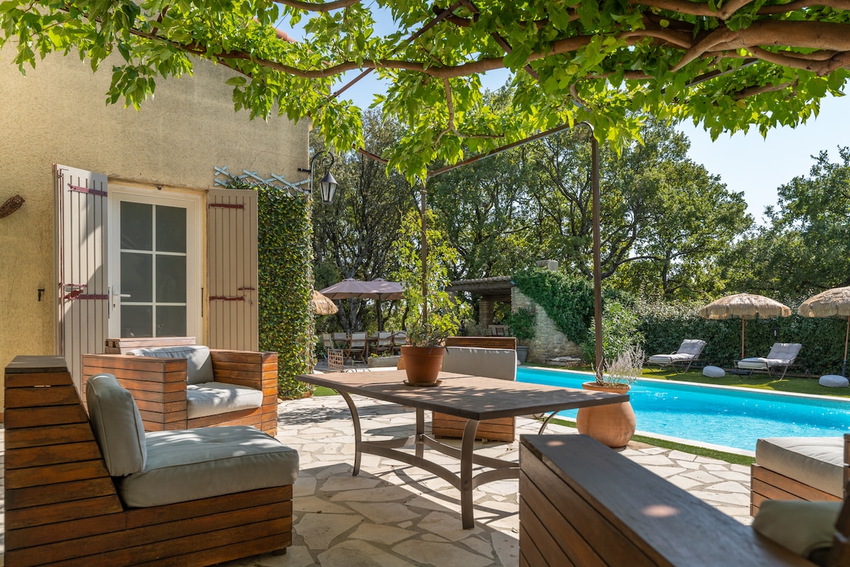 A shaded outdoor seating area features comfortable wooden furniture arranged around a stone table. The pool glimmers in the background, framed by lush greenery and lounge chairs. Sunlight filters through the vines overhead, creating a relaxing atmosphere.