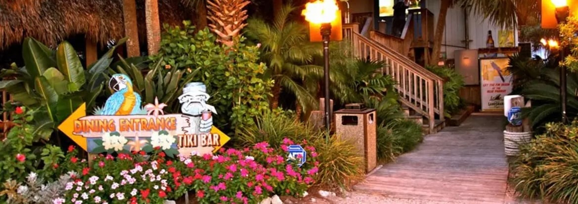 A path leading to a tiki bar entrance is framed by tropical greenery and vibrant pink flowers. Tiki torches provide ambient lighting, creating a welcoming atmosphere. The scene is complemented by palm trees and a nearby trash receptacle, ensuring a neat environment.