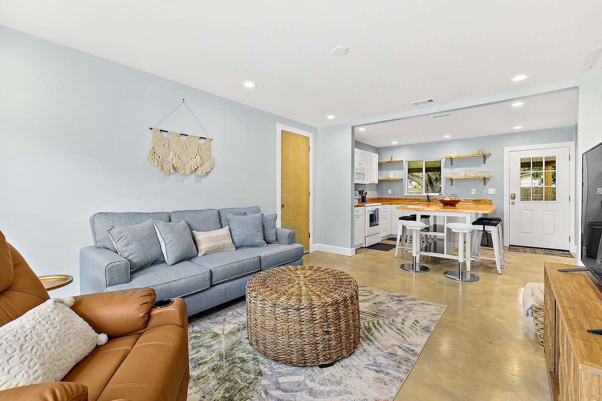 A bright living area features a comfortable sofa and a woven coffee table. Natural light enters through windows, illuminating the open kitchen with modern appliances and a breakfast bar. The space is designed for relaxation and socializing, with a tasteful blend of textures and colors.