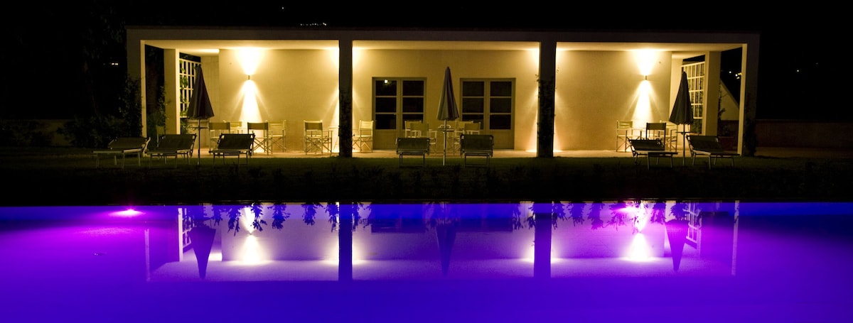 The pool area is softly illuminated at night, showcasing a serene purple hue reflecting on the water's surface. Comfortable lounge chairs and an umbrella are visible, set against the backdrop of a well-lit building featuring large windows.