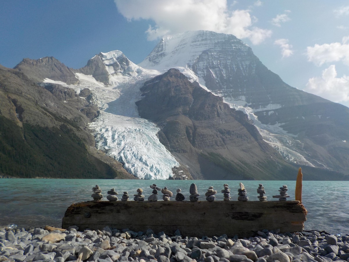 A serene landscape features a clear blue lake in the foreground, bordered by smooth stones and a weathered log. Towering mountains, adorned with snow and glaciers, rise majestically in the background under a partly cloudy sky.