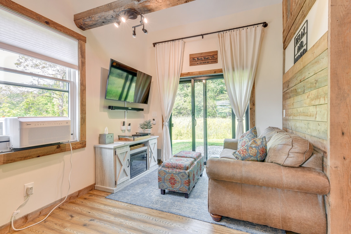 A welcoming living area features a comfortable couch and decorative ottoman, complemented by wood accents. A mounted flat-screen TV and electric fireplace provide modern amenities. Natural light filters through large windows, revealing views of the surrounding greenery, while soft curtains enhance the cozy atmosphere.