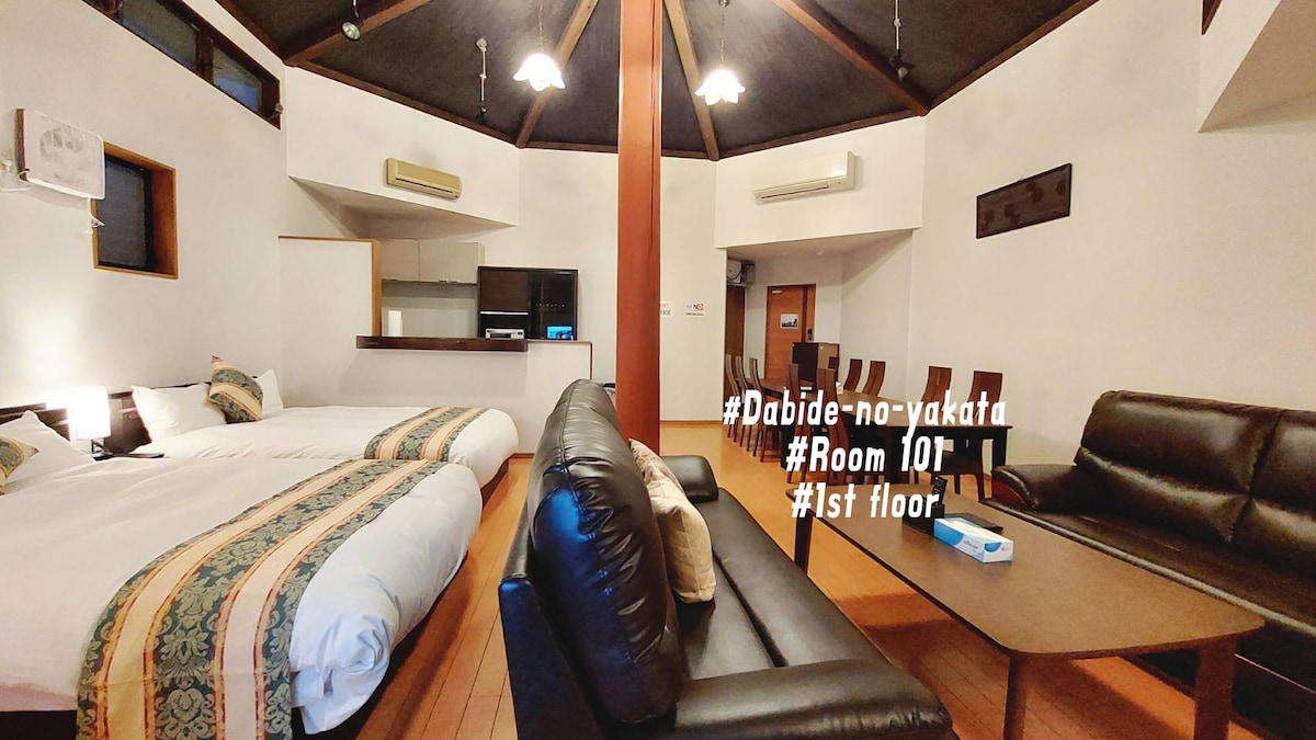A spacious living area features two double-sized beds and a comfortable sofa set. A dining table is positioned nearby. Natural light enters through windows, highlighting the vaulted ceiling and wooden accents. The kitchenette is visible in the background, equipped with modern appliances.