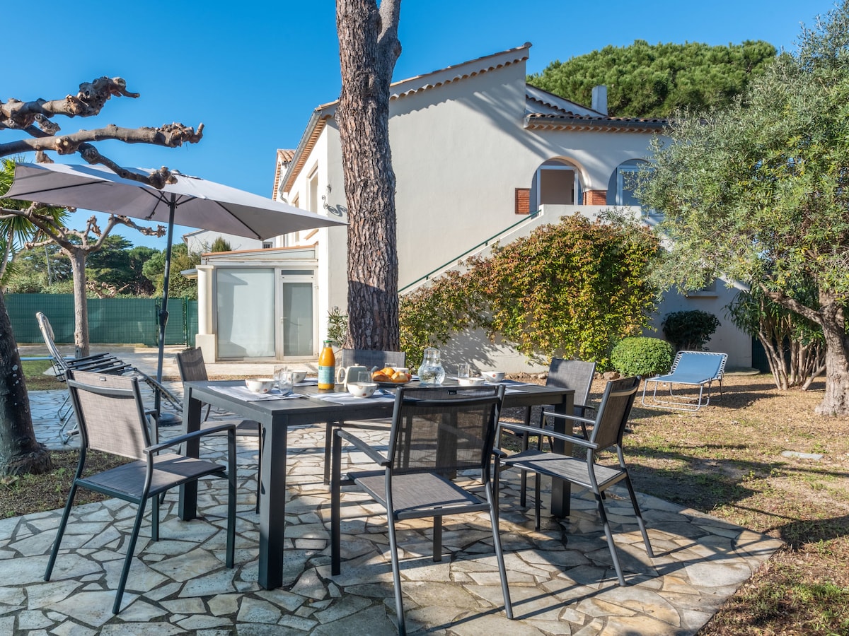 An outdoor dining area features a table surrounded by several chairs, set on a stone pathway. A large umbrella provides shade, and breakfast items are displayed on the table. In the background, a three-story house and well-maintained garden are visible, alongside sun loungers.