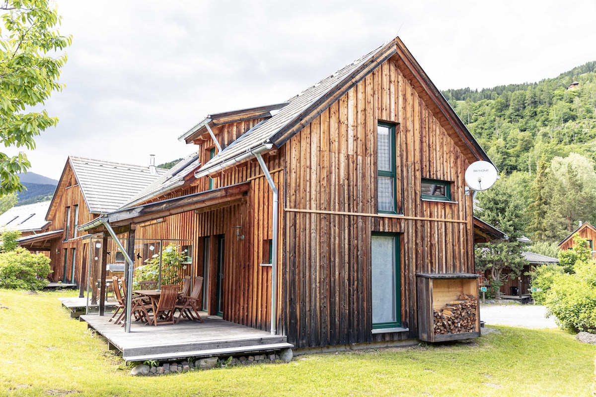 A wooden vacation home is depicted, featuring a large porch with multiple wooden chairs. The exterior includes large windows and is surrounded by greenery. A woodpile is visible beside the entrance. The landscape includes gentle hills in the background under a cloudy sky.
