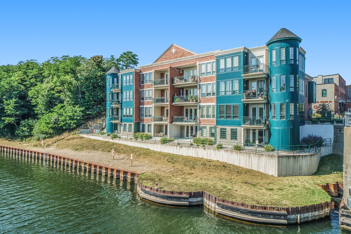 A modern condominium building is situated along the Manistee Channel, featuring a mix of brick and teal-colored exteriors. Multiple balconies are visible, offering views of the water, while greenery lines the shore. A grassy area leads down to the water's edge with a wooden retaining wall.