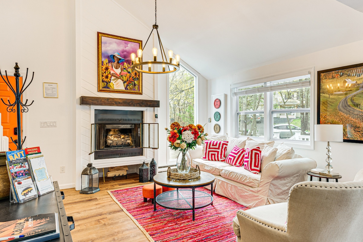 A welcoming living space is presented with a white sofa adorned with colorful pillows. A round coffee table is centered on a vibrant area rug, while a gas fireplace is framed by wall art and decorative elements, allowing natural light to illuminate the room through large windows.