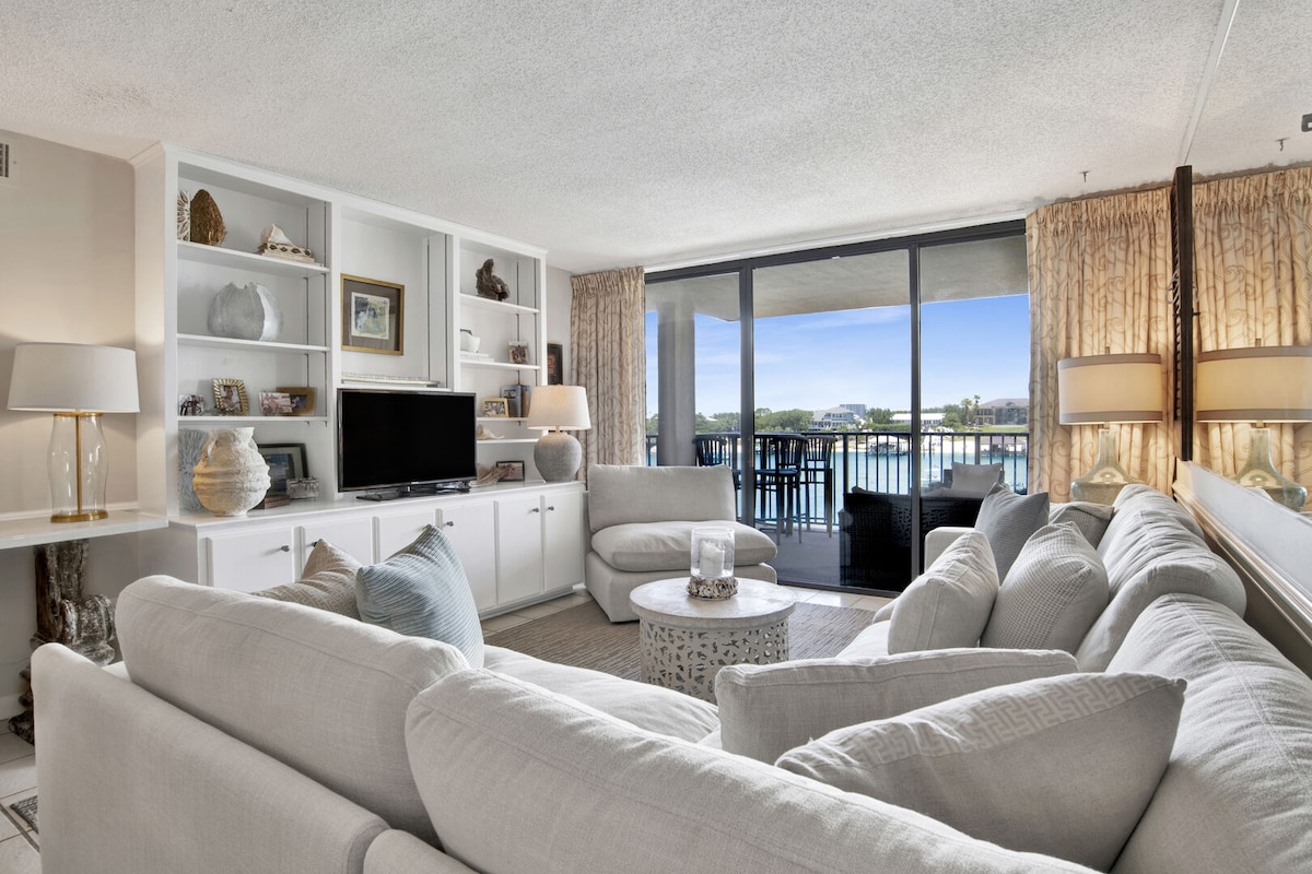 A spacious living area is seen with large, soft sofas arranged around a light-colored coffee table. A flat-screen TV is mounted on built-in shelves, while large glass doors provide natural light and an open view to the balcony overlooking the Ole River.