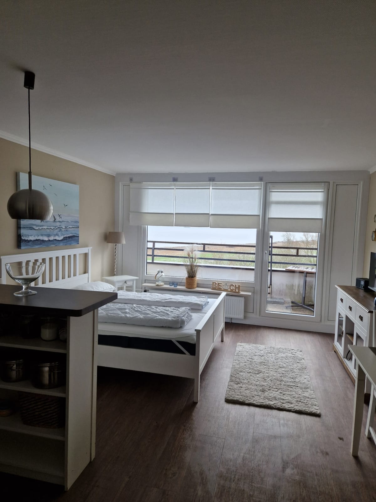 The image displays a bright bedroom featuring a spacious bed positioned near a large window. Natural light floods the room, highlighting a cozy rug and minimalist decor. A small shelf with storage is visible, and a balcony can be seen through the window.
