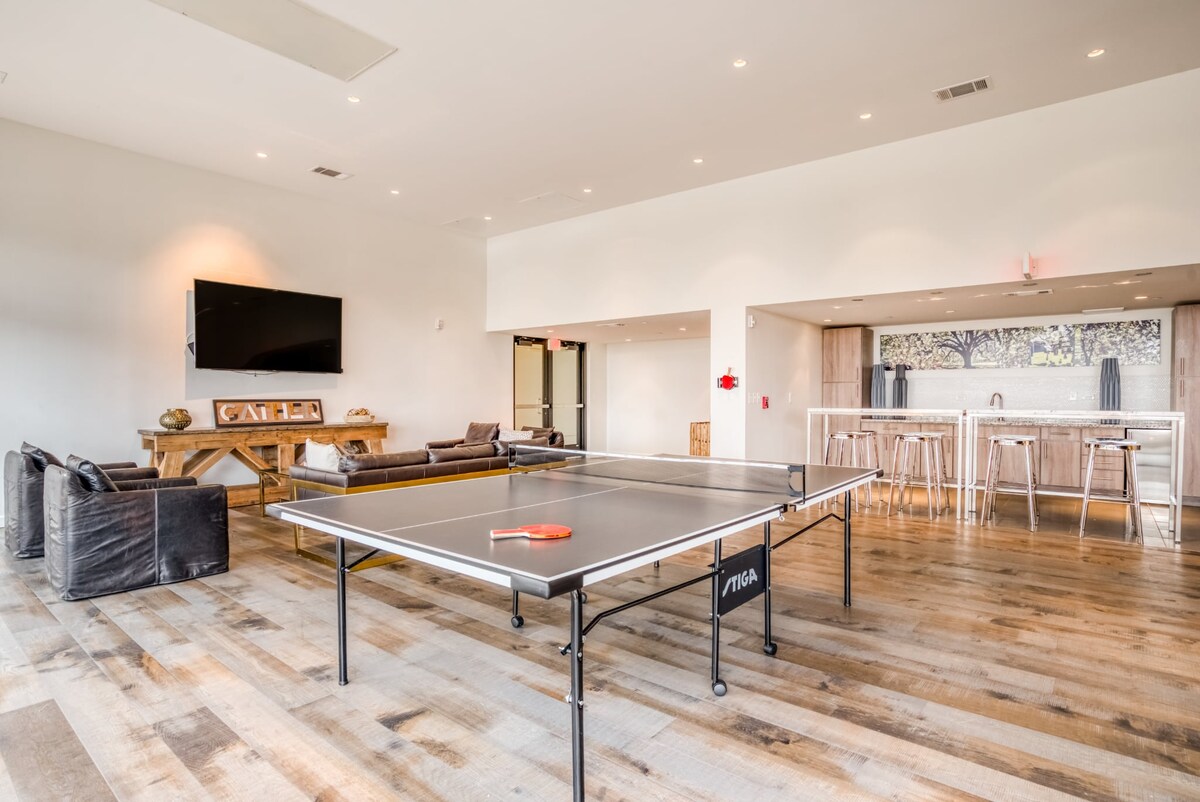 A recreational area is featured, showcasing a ping pong table at the center. Soft seating arrangements, including a sofa and chairs, are positioned nearby. A wall-mounted TV is visible, and a kitchen counter with bar stools is present in the background.