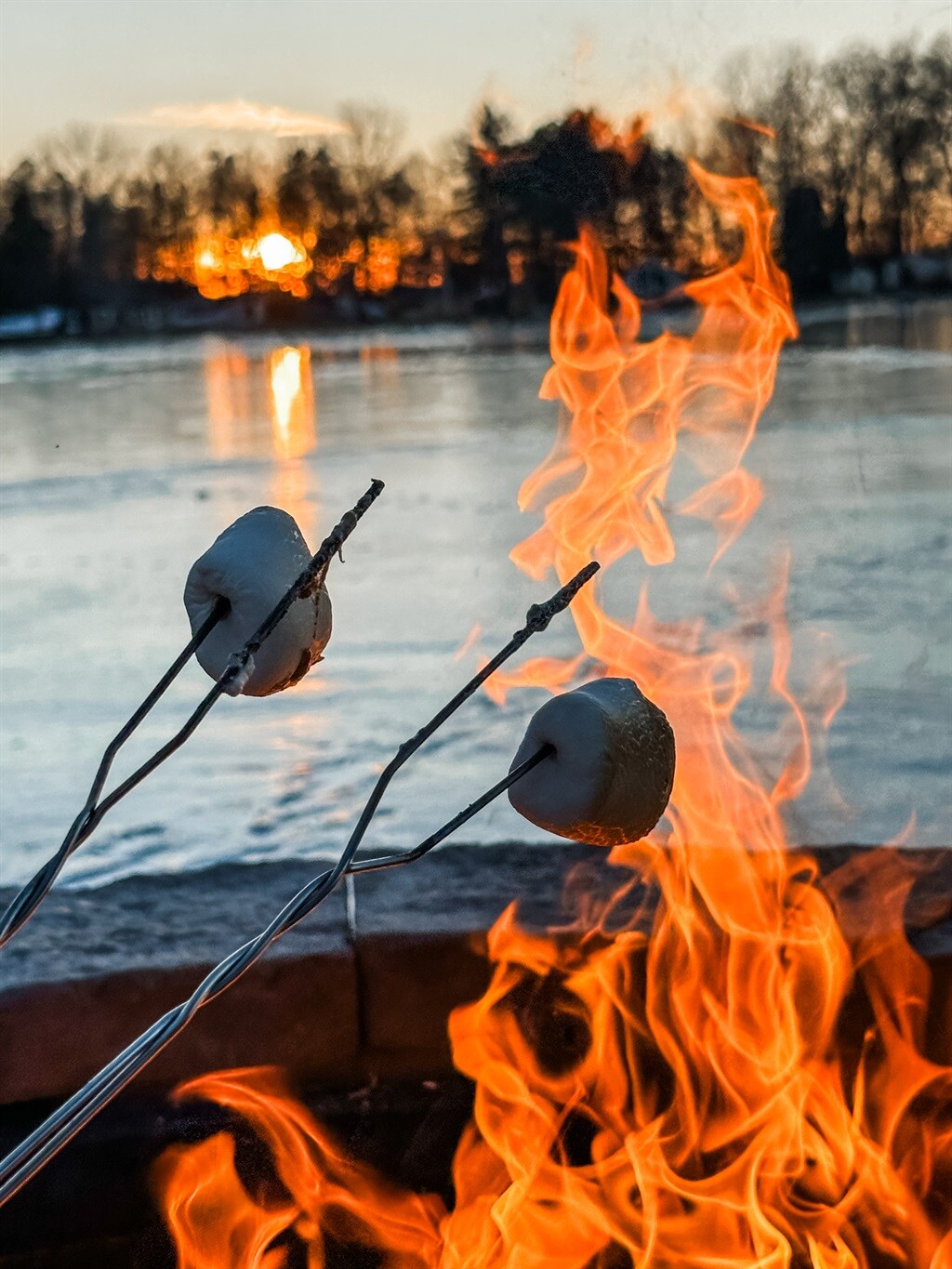 Two skewers hold marshmallows above a vibrant fire pit. Flames flicker intensely against a backdrop of a serene lake reflecting warm sunset hues. Trees are silhouetted against the sky, which displays dusk colors transitioning from orange to deep blue.