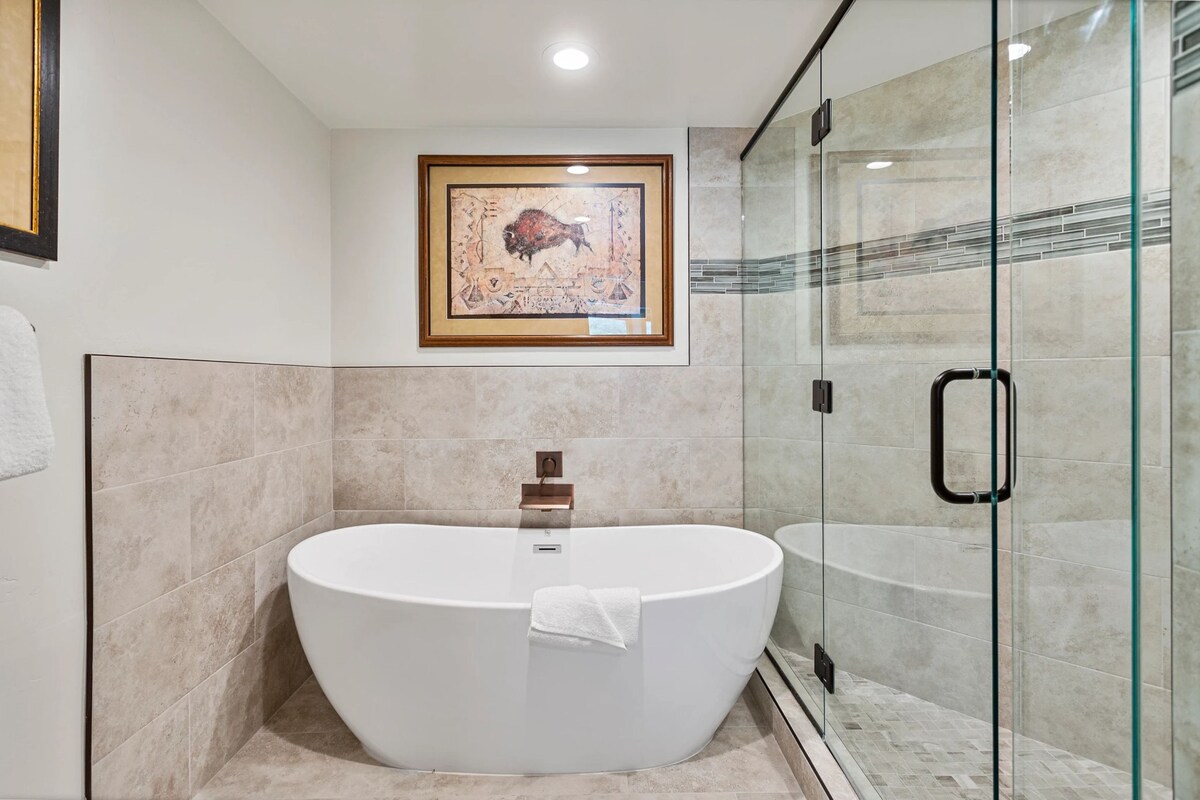 The bathroom features a modern freestanding tub alongside a glass-enclosed walk-in shower. Natural stone tiles create a sleek backdrop, accented by decorative artwork above the tub. Well-lit recessed lighting enhances the spacious feel, while textured stone completes the overall design.