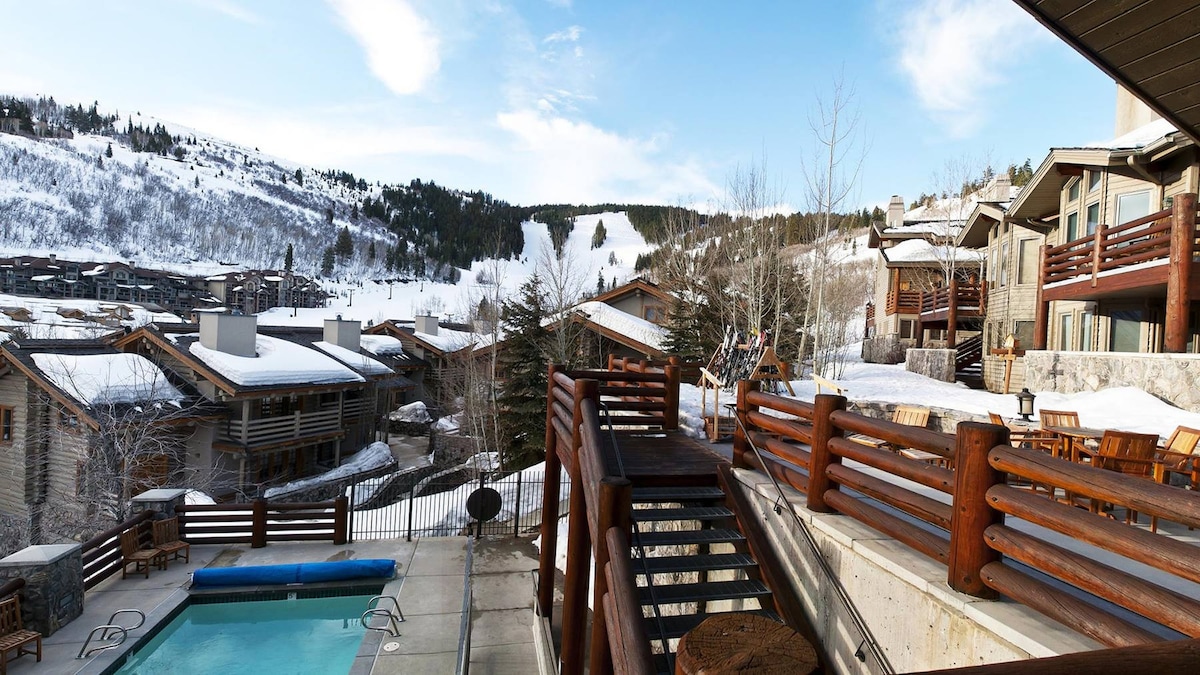 A scenic outdoor area features a heated swimming pool surrounded by snow-covered landscapes. Rustic wooden railings and lounge chairs create a comfortable setting. Nearby, ski slopes are visible in the distance, framed by tree-covered hills under a clear blue sky.