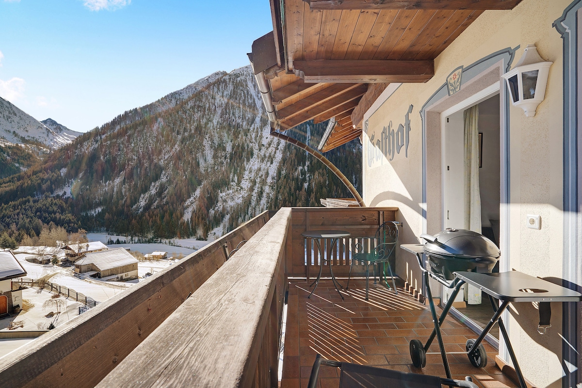 A private balcony features a table and chairs, offering panoramic views of snow-capped mountains and lush valleys. Natural light floods the area, with wooden beams accentuating the warm atmosphere. A barbecue grill is positioned nearby, ready for outdoor cooking.
