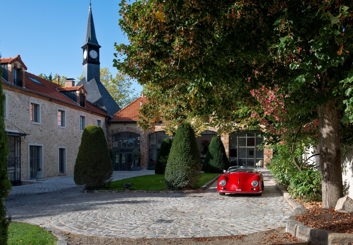 A cobblestone driveway welcomes guests, framed by lush greenery and neatly trimmed bushes. A classic red sports car is parked prominently, with a charming clock tower in the background. The surrounding architecture features a mix of stone and modern elements, adding character to the entrance.