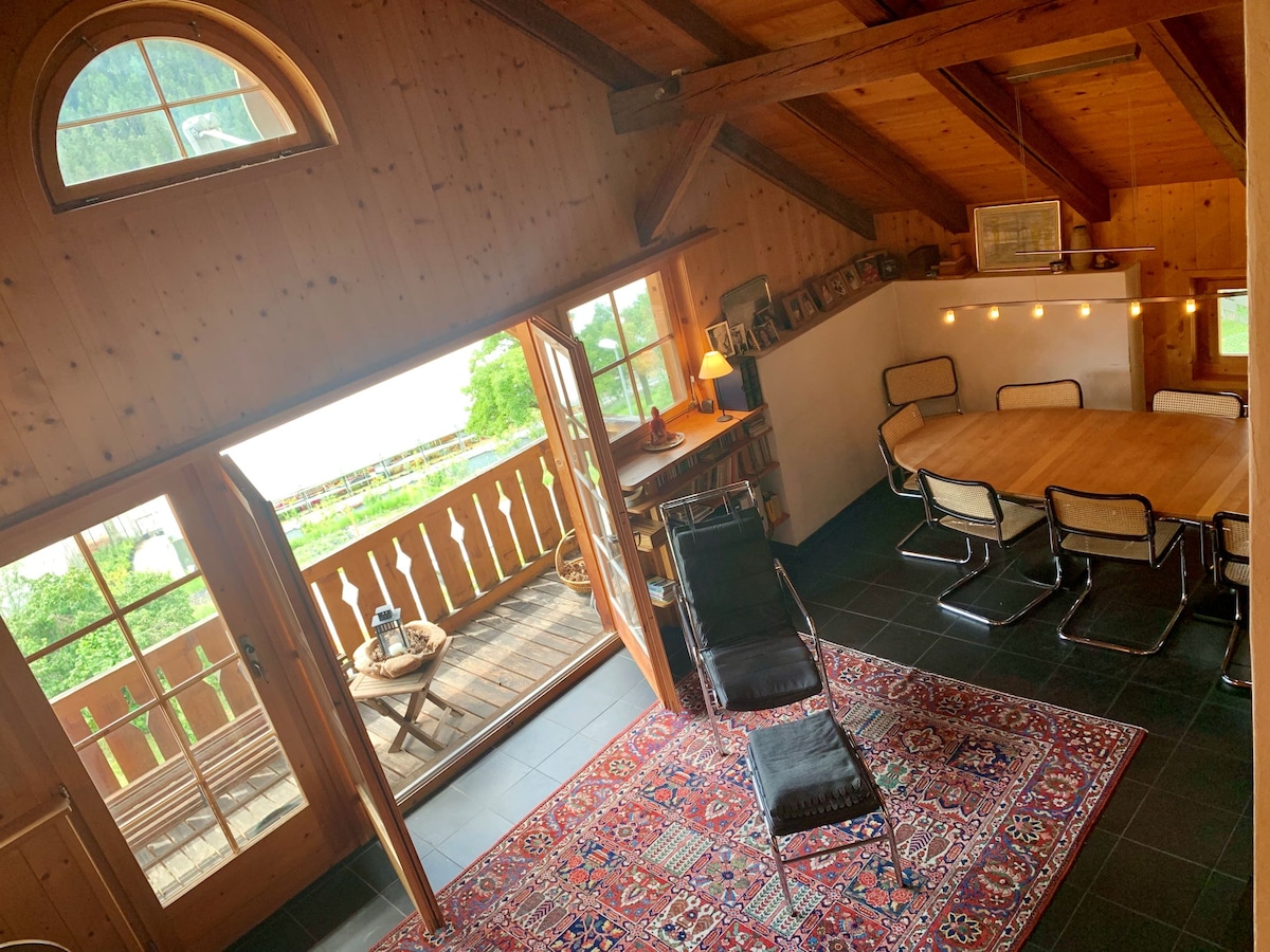 An inviting living space features wooden beams and ample natural light from large windows. A wooden dining table is surrounded by chairs, while a comfortable lounge chair rests on a patterned rug. A balcony extends outdoors, providing views of the surrounding greenery.