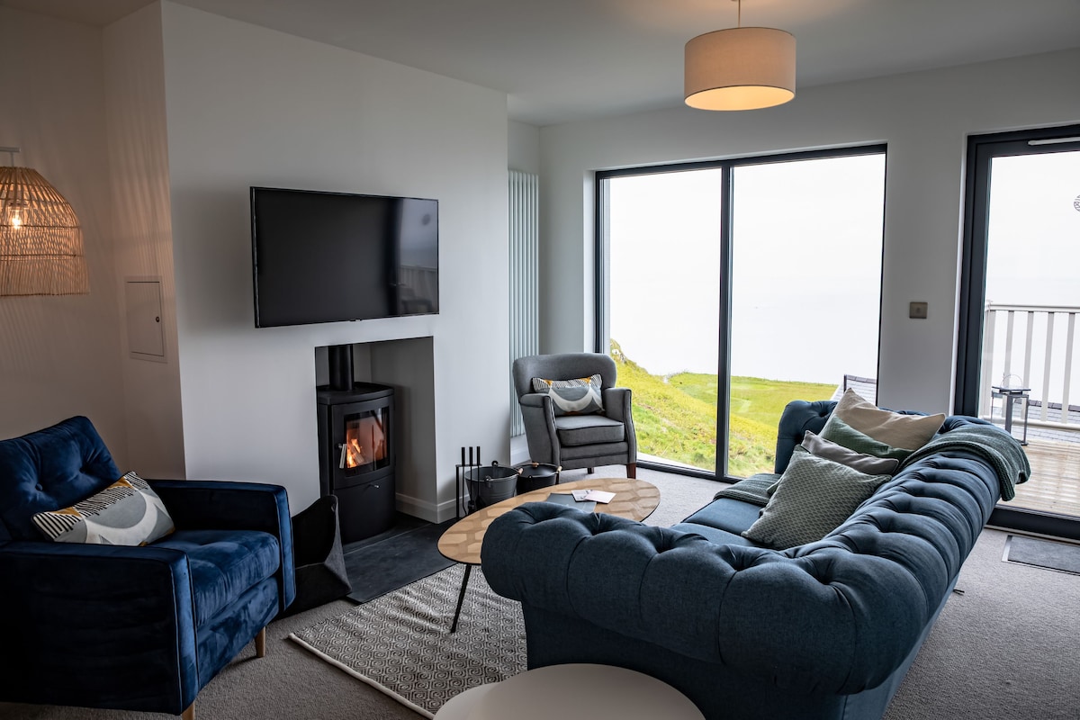 A light-filled living area features a plush blue sofa and a cozy armchair, with large windows offering views of the expansive green landscape. A modern coffee table sits centrally, while a sleek TV and a stylish fireplace complete the inviting atmosphere.