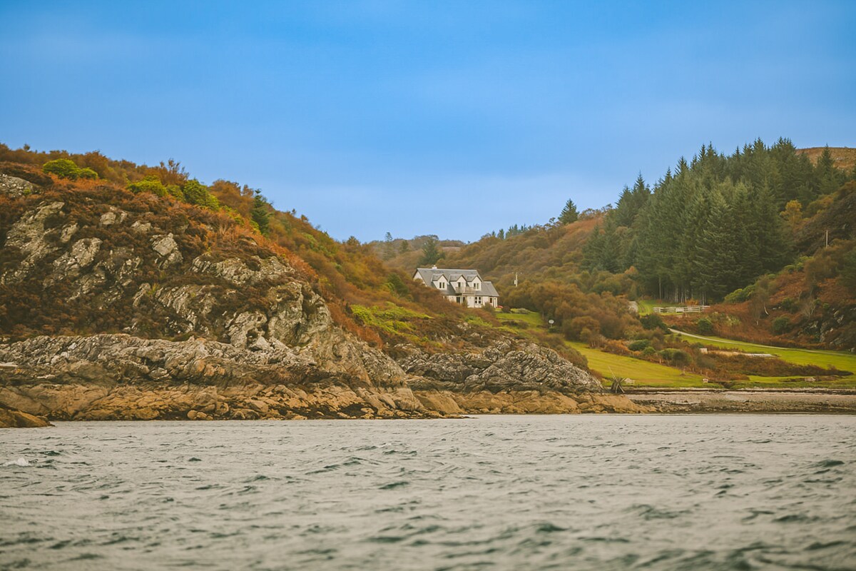 The holiday house is positioned on elevated terrain overlooking a serene bay. Natural rocky formations and lush greenery frame the property, providing a tranquil coastal setting. The structure features a light exterior that complements the surrounding landscape under a soft blue sky.