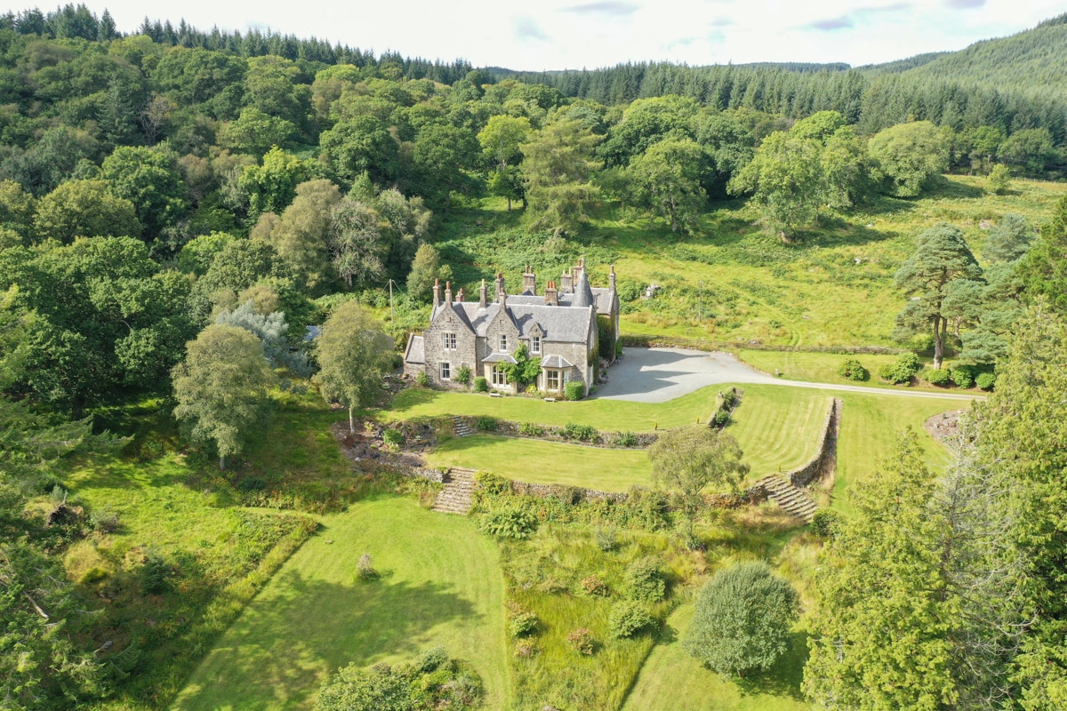 The exterior of Tarbert Country Manor is surrounded by lush greenery and trees. The stone manor features a traditional design with multiple chimneys and a well-maintained garden. A gravel driveway leads to the entrance, providing ample space for vehicles against a backdrop of natural beauty.