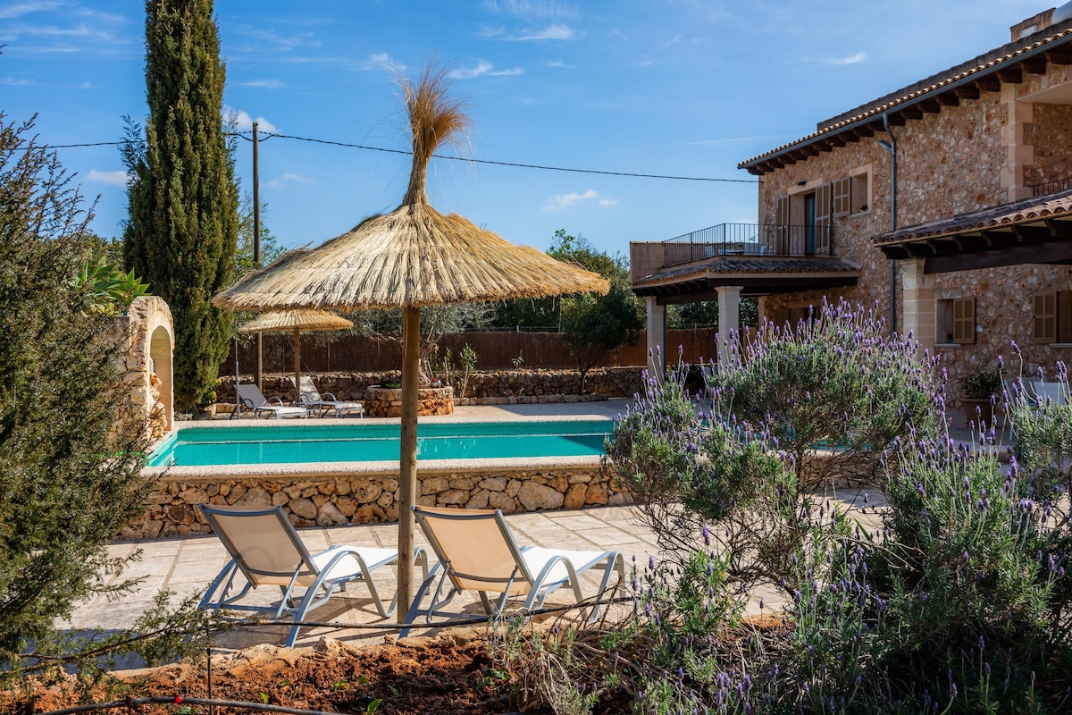 A shared outdoor area features a swimming pool surrounded by a stone terrace. Sun loungers are arranged underneath a thatched umbrella, offering shade. Lush greenery and lavender plants are visible in the garden, enhancing the tranquil atmosphere.