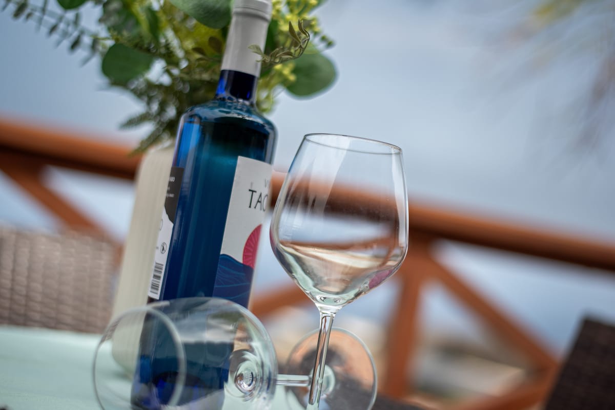 A blue wine bottle rests upright on a table, next to an elegantly shaped wine glass placed on its side. A green plant in the background adds a touch of nature, while the distant view of the sea can be subtly seen.