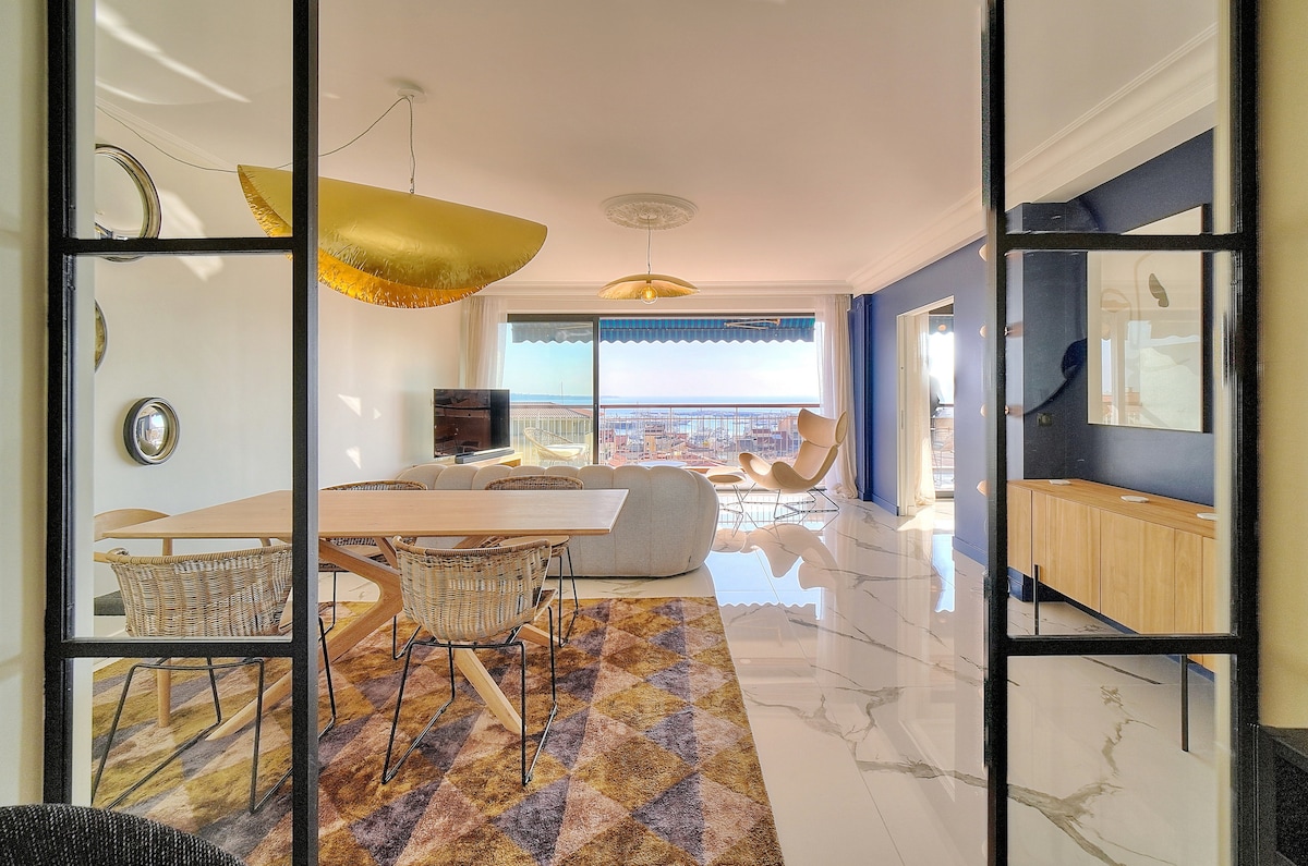 The interior features an open living space with large windows allowing natural light. A dining table is placed centrally, surrounded by four woven chairs. A comfortable sofa and an accent chair are positioned in the corner, with a flat-screen TV mounted on a wooden cabinet. Elegant decorative elements adorn the walls.