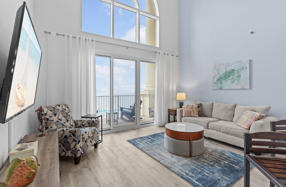 A spacious living area is highlighted by large windows allowing ample natural light. A comfortable sofa is paired with a patterned armchair. A round coffee table is centered on an area rug, while sliding glass doors provide access to a private balcony with views of the surroundings.