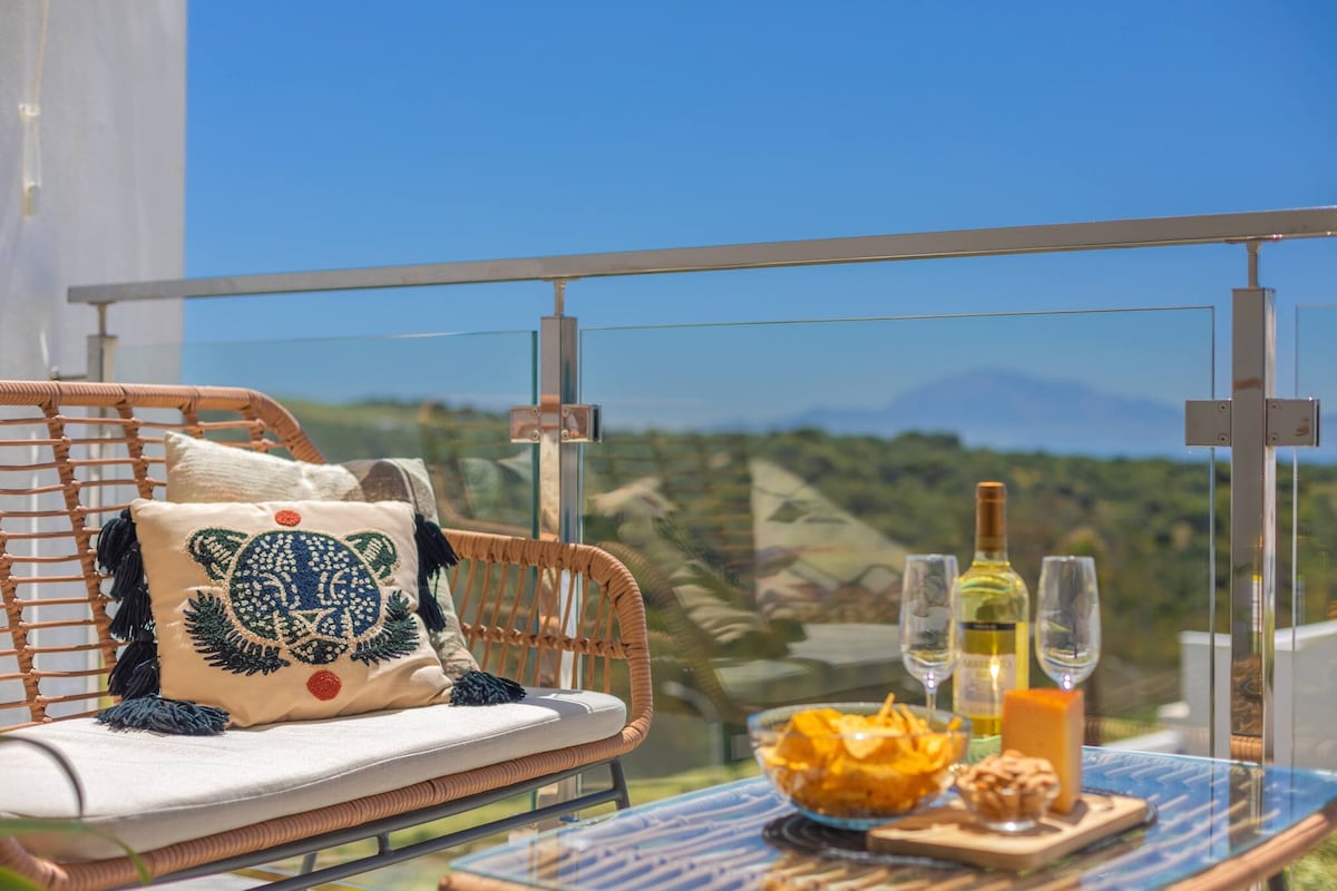 A comfortable seating area is highlighted on the balcony, featuring a rattan sofa adorned with a decorative cushion. A glass table holds a bottle of wine, two glasses, and a plate of snacks, while a picturesque landscape is visible in the background.