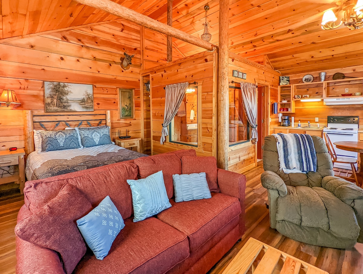 A cozy cabin interior features wooden paneling and a spacious bed draped in blue bedding. A red sofa with accent pillows invites relaxation, while a green armchair adds comfort. The kitchenette is visible in the background, complemented by a small dining area.