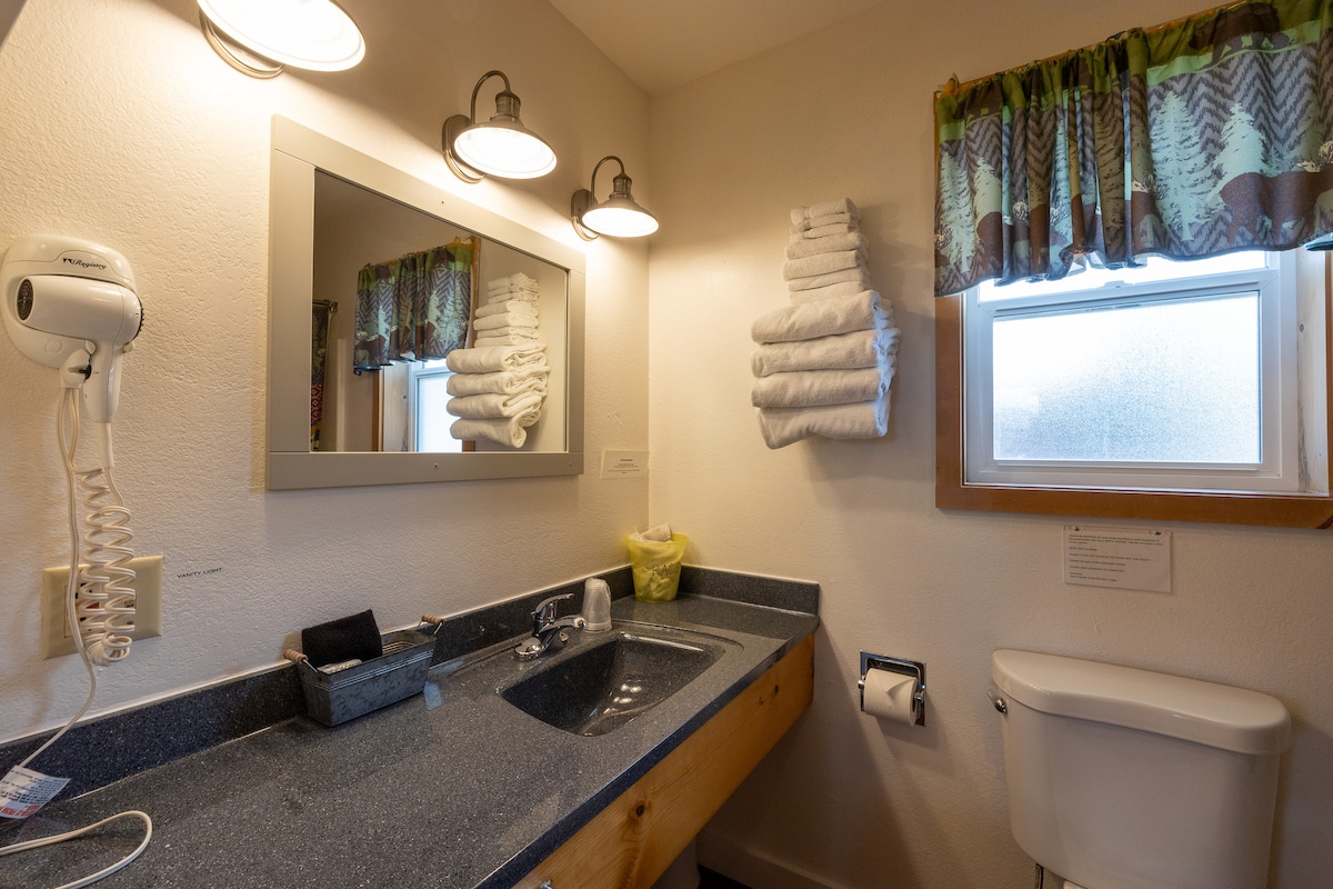 A spacious bathroom features a countertop with a sink and a large mirror above. Fresh white towels are neatly arranged on the counter and hung on a rack. A window with a patterned curtain allows natural light, while a toilet is positioned beside the sink area.