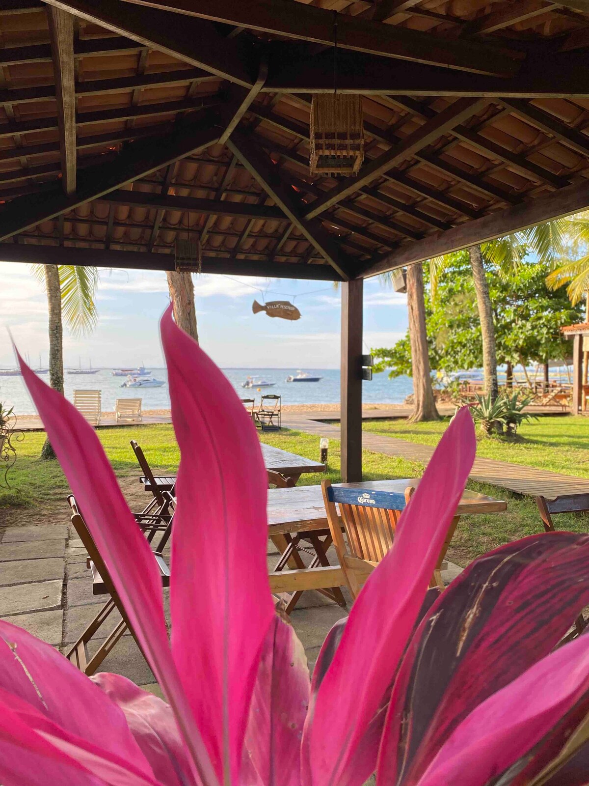 A vibrant pink leaf extends into the foreground, contrasting with the natural surroundings. The view features an outdoor seating area under a wooden roof, overlooking a tranquil beach scene with boats in the distance. Palm trees and greenery add to the serene atmosphere.