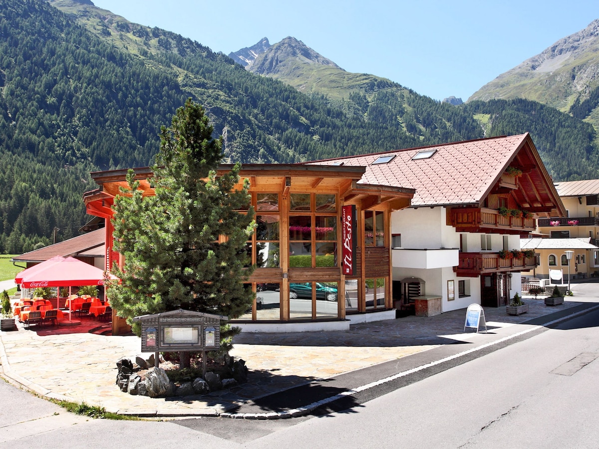 A welcoming building set in a scenic mountain area, featuring a wooden exterior with large windows. Surrounding greenery enhances the setting, and a patio area with red umbrellas provides outdoor seating. The clean pathway leads to the entrance, inviting guests into a comfortable space.