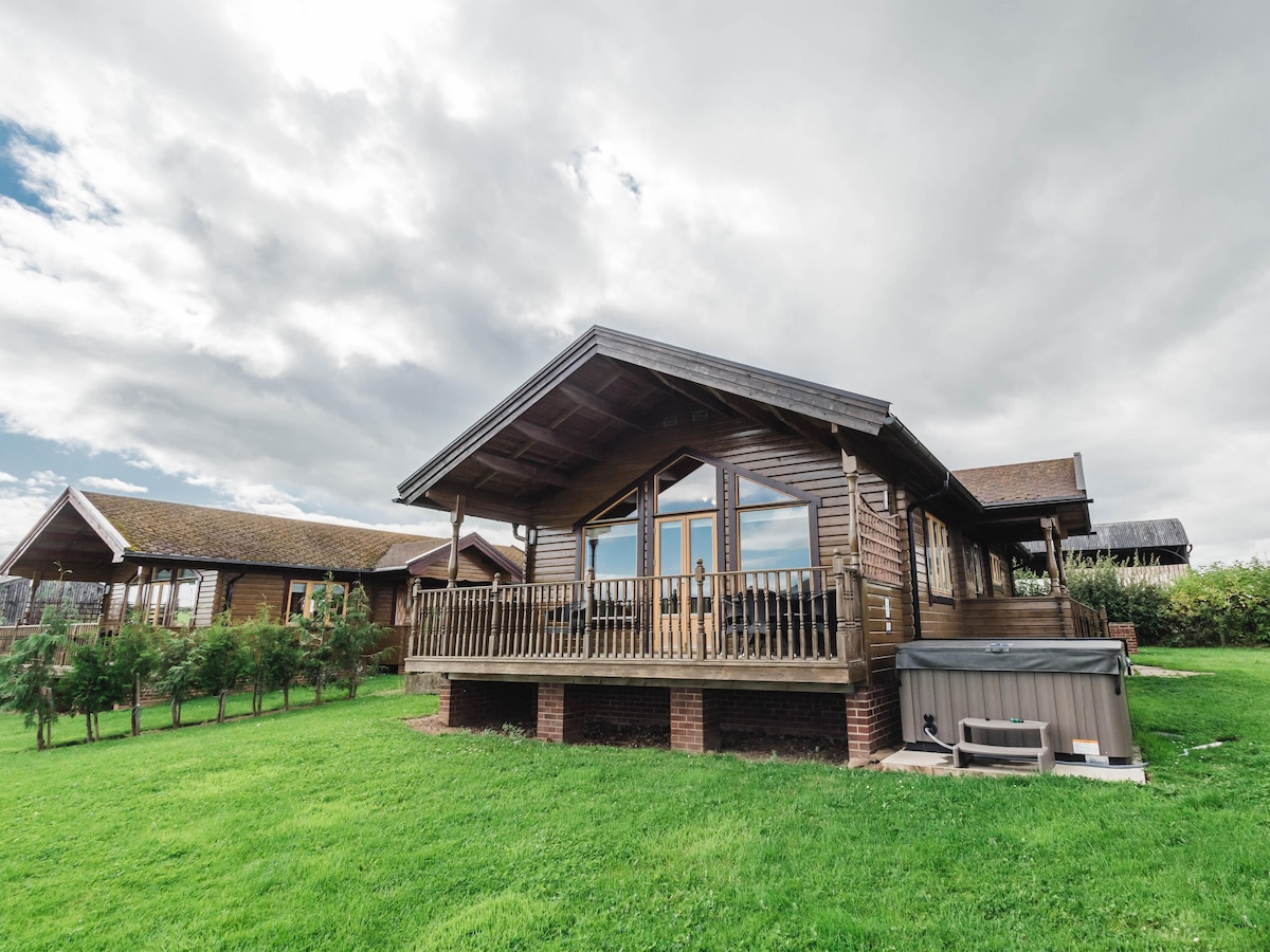 A wooden lodge is set against a backdrop of cloudy skies, featuring large front windows that offer views of the surrounding greenery. A spacious deck with railing extends from the lodge, and a hot tub is located on the ground level nearby.