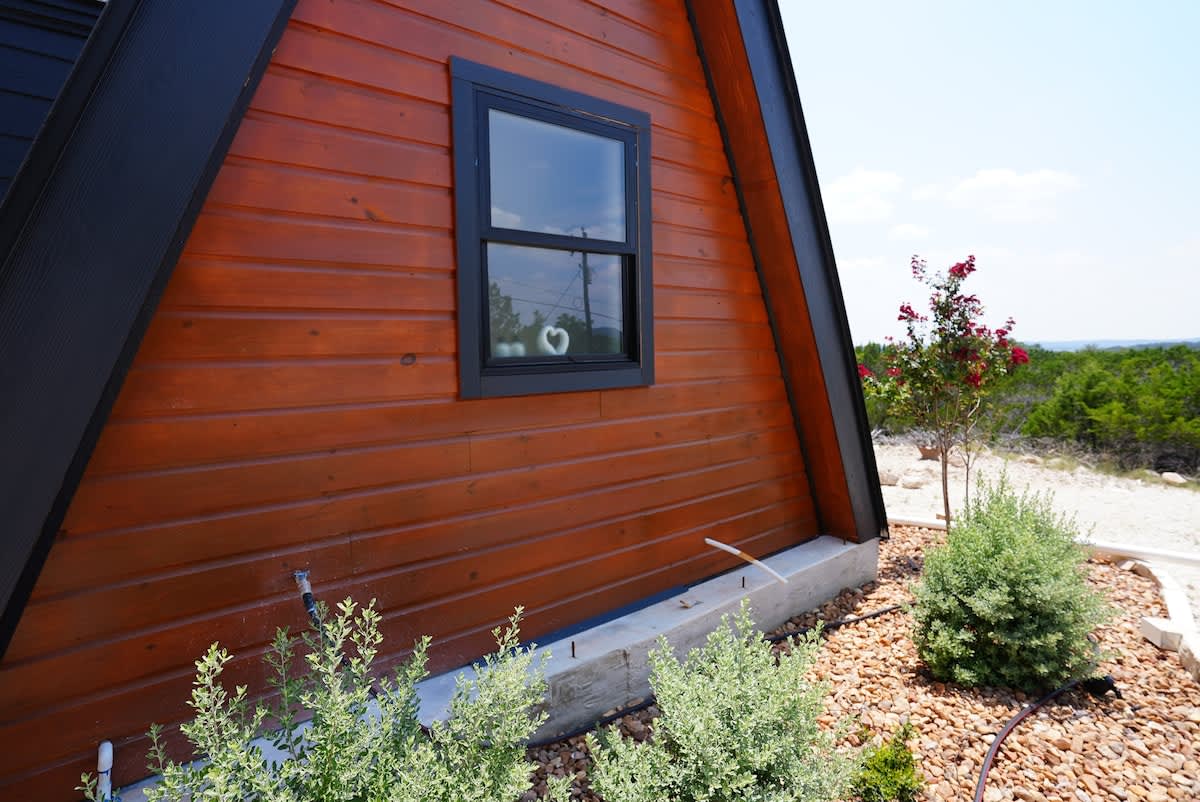 The exterior of a charming A-frame cabin is highlighted, showcasing rich wooden paneling and a large window framed in black. Lush greenery and decorative stones surround the base, complemented by a blooming shrub nearby.