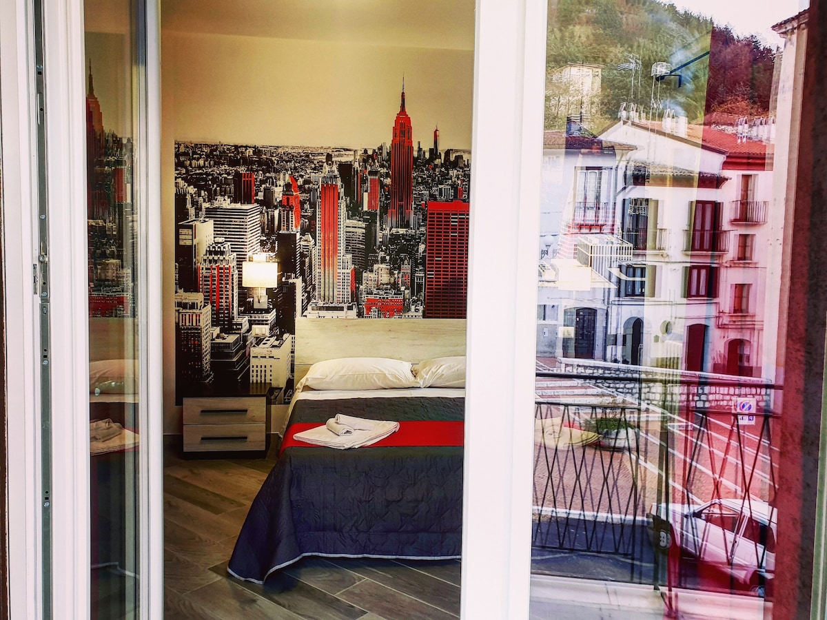A stylish room with an urban mural features a spacious bed adorned with a dark cover. Natural light filters through the balcony doors, illuminating the modern decor. The scene outside showcases a glimpse of the surrounding architecture and greenery.