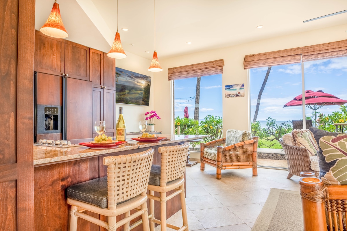 The inviting kitchen area features a bar height countertop with a seating arrangement for four. Two glasses and a bottle sit on the counter, complemented by tasteful décor. Large windows frame views of the lush landscape and ocean beyond, while natural light enhances the warm interior.