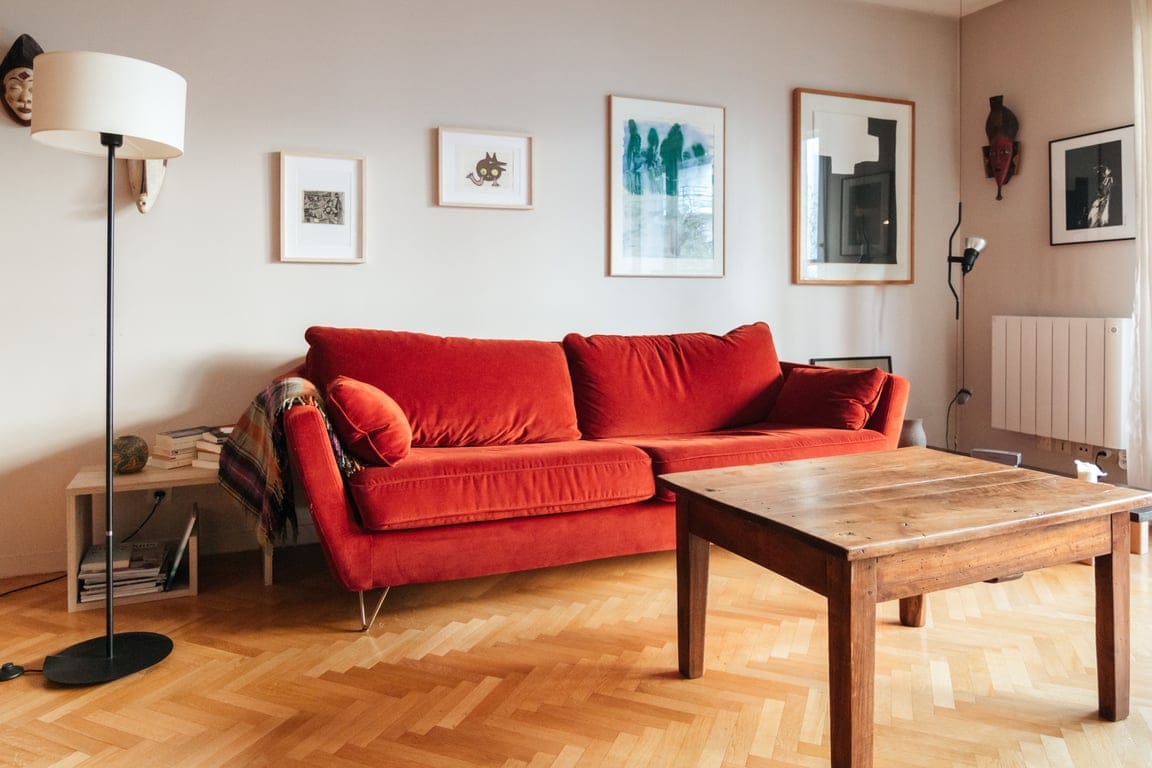 A vibrant red sofa is positioned beside a wooden coffee table on a herringbone-patterned floor. Framed artwork decorates the light-colored walls, while a stylish lamp stands nearby, contributing to a well-lit and inviting living area.