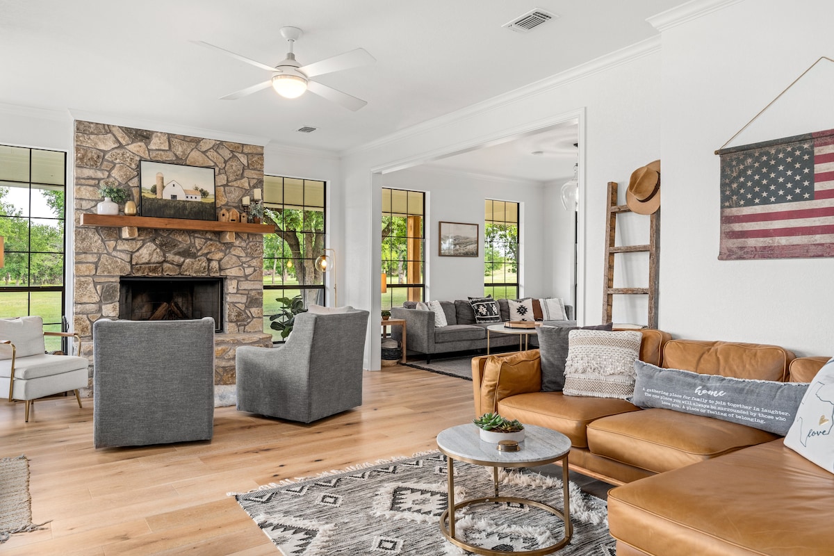 A spacious living area is highlighted by a stone fireplace, accented by comfortable seating arrangements. Soft, neutral tones create a relaxing environment, with large windows offering views of the surrounding nature. Warm wood flooring complements the modern decor, inviting conversation and relaxation.