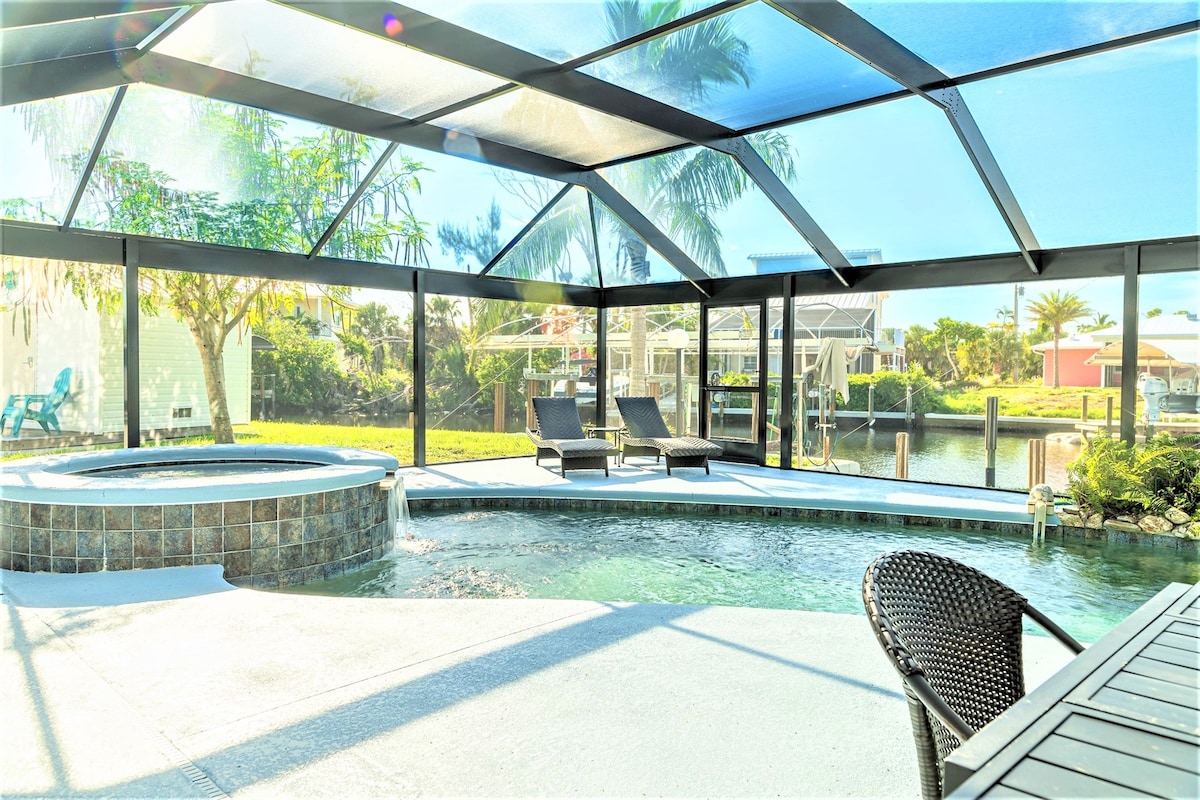 A screened-in outdoor space features a private saltwater pool and adjacent hot tub, surrounded by a patio area. Lounge chairs are positioned for relaxation, while views of the canal and nearby homes are visible through the mesh enclosure. Lush greenery adds a natural touch.