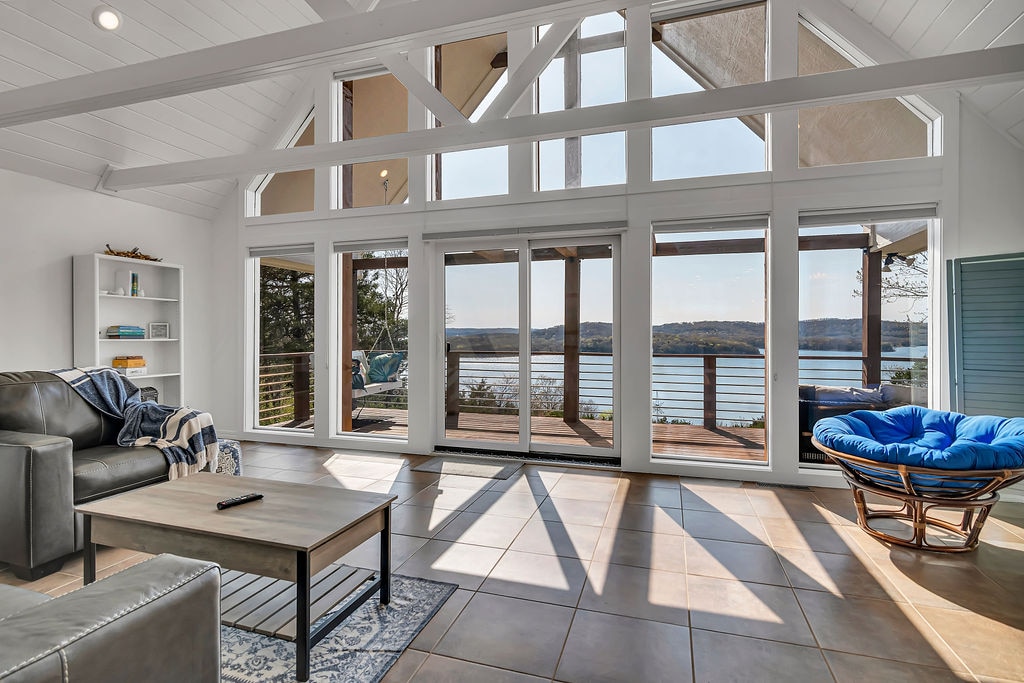 A spacious living area features tall windows that provide expansive views of the lake. Natural light floods the room, highlighting a comfortable sofa and a round chair. A coffee table sits on tiled flooring, creating a warm and welcoming environment.