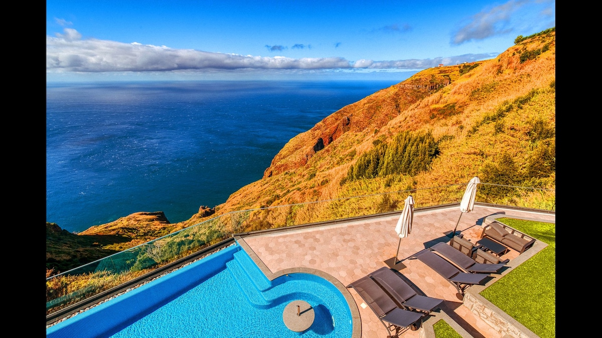A serene outdoor area features a spacious infinity pool overlooking a stunning coastal view. Sun loungers arranged around the pool offer comfort, complemented by two cantilever umbrellas. The rugged landscape of cliffs and the expansive Atlantic Ocean are visible in the background.