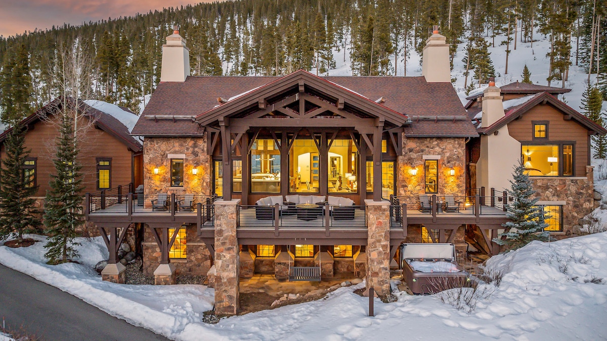 The exterior of Switchback Lodge is highlighted by rustic stone and wood elements, framed by snow-covered trees. An expansive covered deck is visible, featuring several outdoor seating areas, with warm lights glowing from the windows, inviting guests to enjoy the serene mountain setting.
