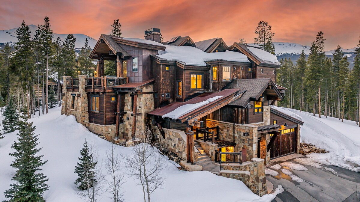 The residence is nestled among snow-covered trees, featuring a combination of wooden and stone exterior elements. The sloped rooflines and multiple levels create a distinctive and warm architectural presence against a backdrop of a colorful sunset sky.