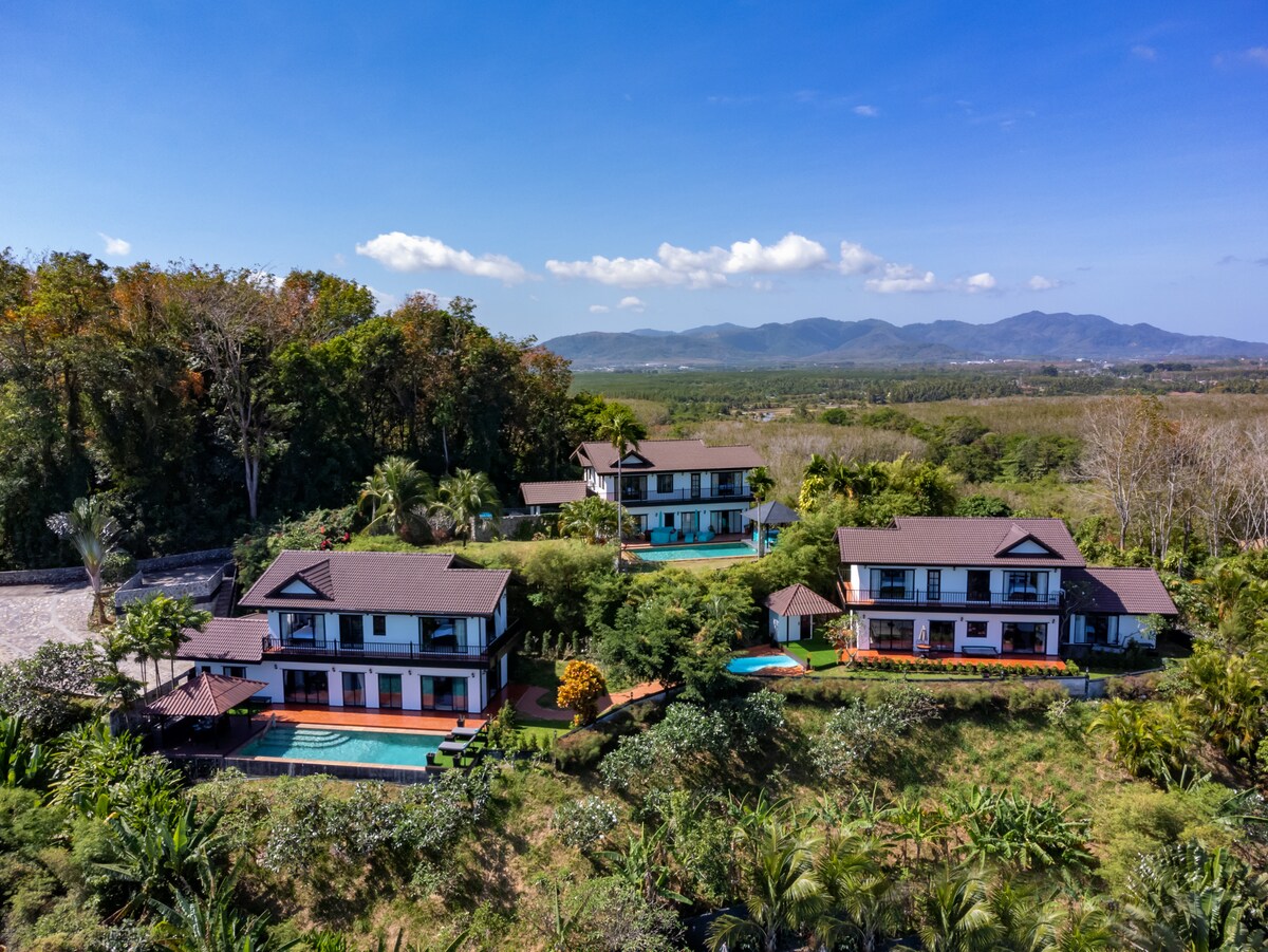 An aerial view of a tranquil villa complex is displayed, surrounded by lush greenery and trees. Each villa features a private pool and is positioned amidst a scenic landscape that includes distant mountains and clear blue skies. Sunlight enhances the natural beauty of the setting.