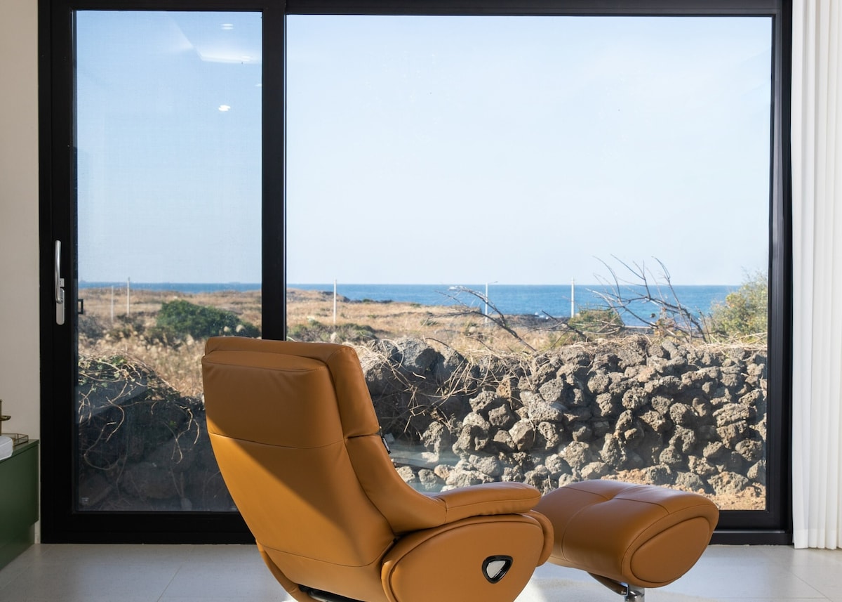 A modern leather recliner is positioned near a large glass window, offering a view of the tranquil ocean and surrounding natural landscape. The scenery includes rocky terrain and stretches of greenery under a clear sky.