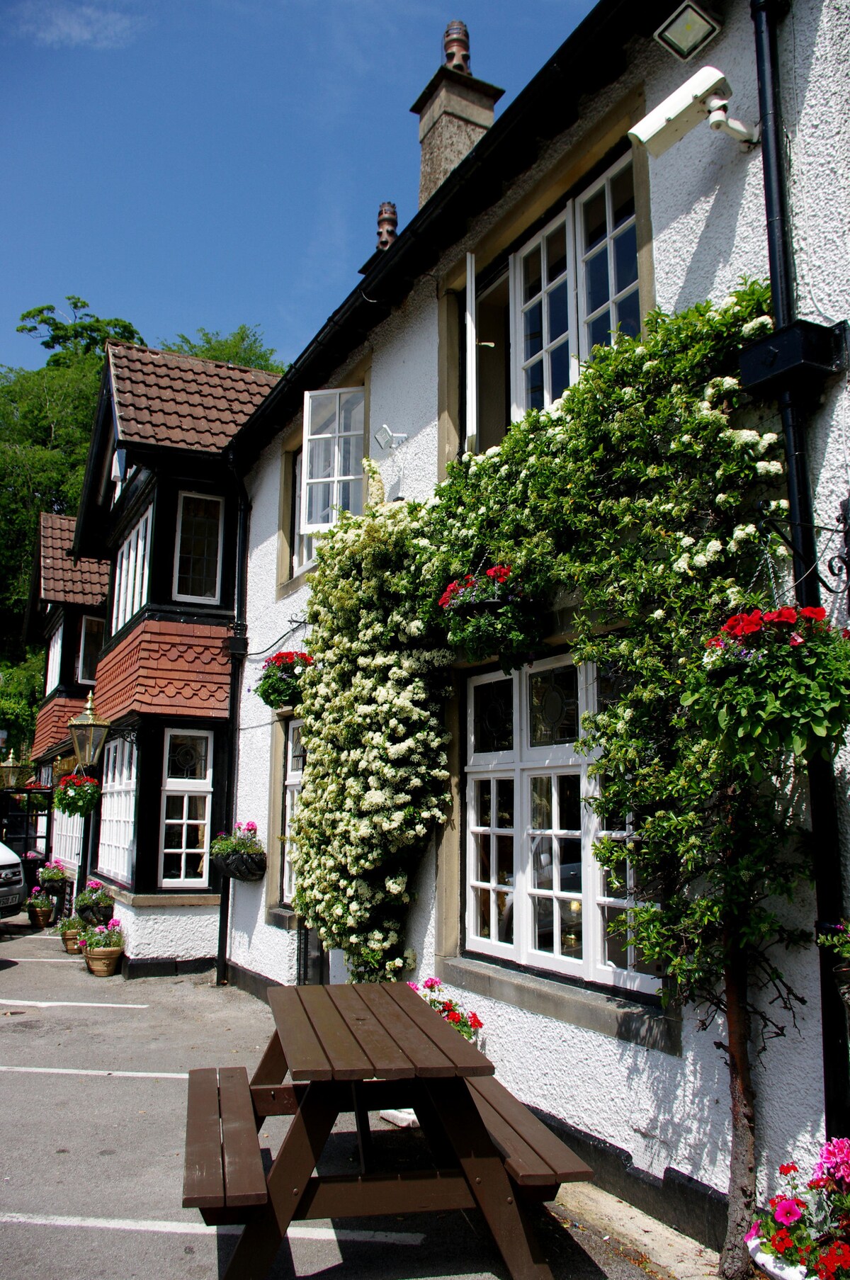 The exterior of a charming building is adorned with vibrant climbing plants and window boxes filled with colorful flowers. A wooden picnic table sits in front, providing an inviting space for guests. The sky is clear, illuminating the structure's character.