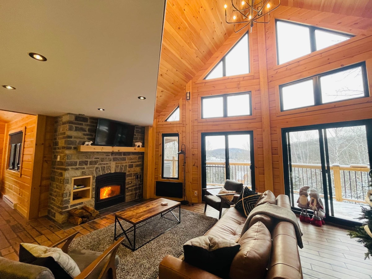The living area features a high cathedral ceiling and large windows, allowing for scenic mountain views. A stone fireplace adds warmth to the space, which is furnished with comfortable leather sofas and a central coffee table. Natural wood finishes enhance the cabin's rustic charm.