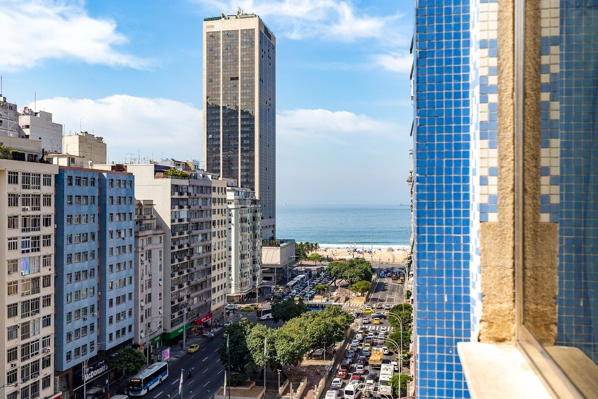 A view from a high window showcases a vast urban landscape with modern buildings lining the street. In the distance, the ocean appears against a backdrop of blue skies, indicating a vibrant coastal atmosphere. Trees dot the area, adding greenery to the scene.